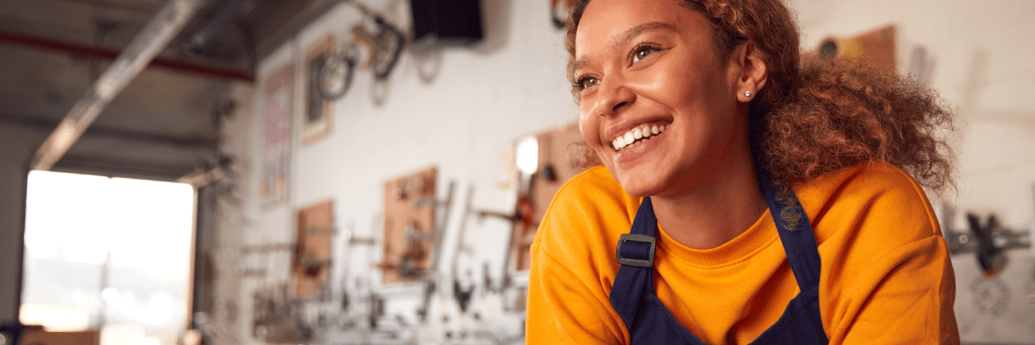 woman in a workshop wearing an orange jumper and a black apron smiling